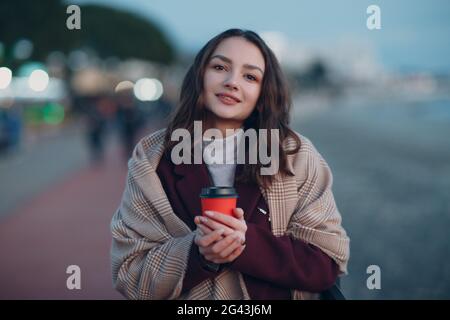 Junge schöne Frau in Mantel und Schal mit Kaffeetasse im Freien. Stockfoto