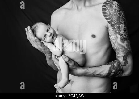 Vater mit Tätowierung am Arm hält sein neugeborenes Baby auf einem schwarz-weißen Hintergrund Stockfoto