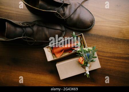 Nahaufnahme einer Fliege für einen Bräutigam mit einem Boutonniere auf einem Karton und Herrenschuhe auf einem braunen Boden. Stockfoto