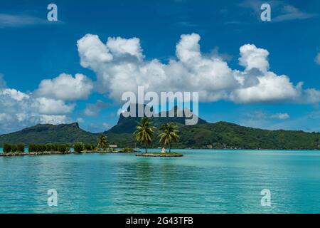 Zwei Palmen und eine Tiki-Skulptur auf einer kleinen Insel am Eingang zum Hafen des Flughafens Bora Bora (BOB) mit dem Berg Otemanu in der Ferne, Bora Bora, Stockfoto