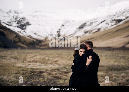 Reiseziel Island Hochzeit. Hochzeitspaar auf einem Hintergrund von verschneiten Bergen. Die Braut und der Bräutigam in schwarzen Mänteln umarmen mich Stockfoto