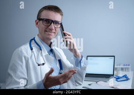 Lächelnder Arzt, der an der Rezeption telefoniert. Physicianr im Gespräch mit seinem Patienten in der Büroklinik am Tisch sitzend mit Laptop. Ger Stockfoto