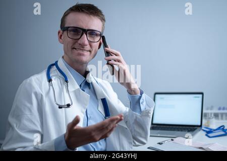 Europäischer Arzt in weißem medizinischen Mantel und Brille konsultiert den Patienten auf dem Handy in der Klinik Büro, sitzen am Tisch mit Laptop. Stockfoto