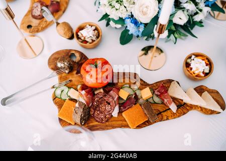 Nahaufnahme eines Schneidebretts mit Tomaten, Wurstscheiben, Prosciutto, Käse und Gurke mit Küchenzange auf einem weißen Tisch Stockfoto