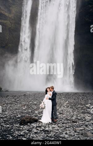 Der Bräutigam umarmt die Braut von hinten. Hochzeitspaar in der Nähe des Skogafoss Wasserfalls. Hochzeit in Island. Stockfoto