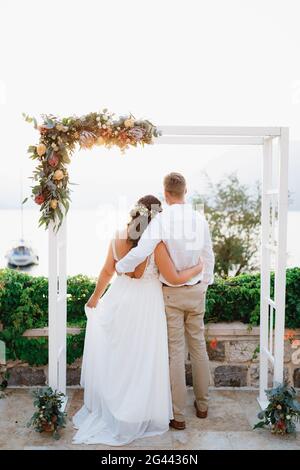 Die Braut und der Bräutigam stehen umarmt unter dem Hochzeitstor und blicken auf das Meer, Rückansicht Stockfoto