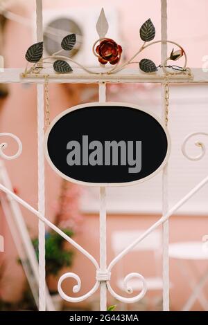 Leeres ovales Schild auf dem Hintergrund eines schönen geschmiedeten Weißer Zaun mit Blumen Stockfoto
