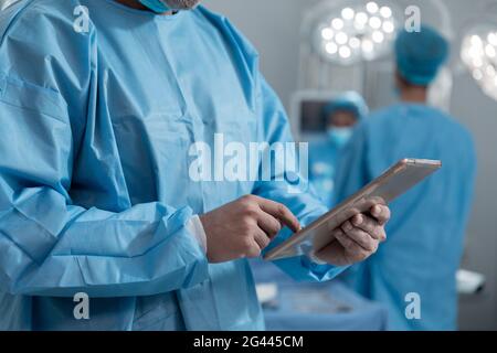 Mittelteil des kaukasischen männlichen Chirurgen, der Schutzkleidung mit einer Tablette trägt Im Operationssaal Stockfoto