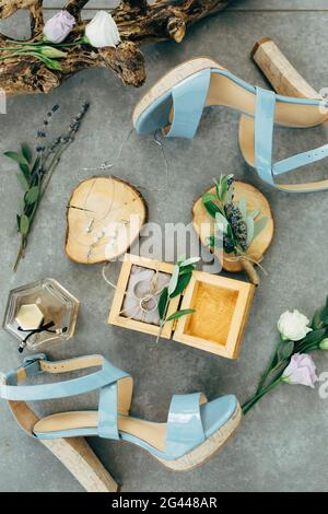 Eheringe liegen in einer Holzkiste, umgeben von Sandalen mit Absätzen, Blumen und Zweigen auf dem Boden Stockfoto