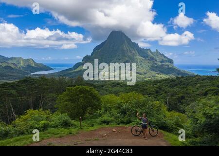 Luftaufnahme einer Frau mit Fahrrad am Aussichtspunkt Belvedere mit Blick auf die Opunohu Bay (links) und Cook's Bay (rechts), Moorea, Windward Islands, French Pol Stockfoto