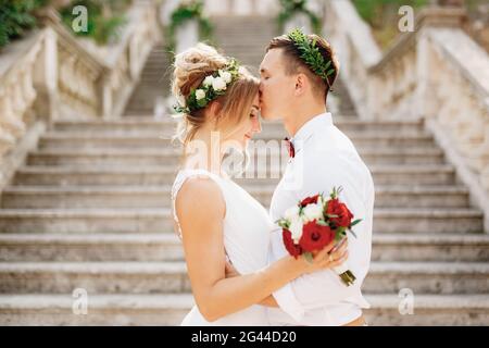 Die Braut und der Bräutigam in Kränzen stehen umarmt und küssend auf der Treppe der Geburt der alten Kirche in Prcanj, Nahaufnahme Stockfoto