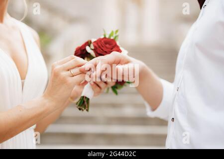 Die Braut legt einen Ehering auf den Finger des Bräutigams Und hält einen Strauß roter und weißer Rosen in sich Ihre Hand Stockfoto