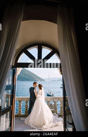 Das Hochzeitspaar steht auf einem Hotelbalkon mit Meerblick, Blick durch ein offenes antikes Fenster. Hochzeitsfoto der feinen Kunst in Montenegro Stockfoto