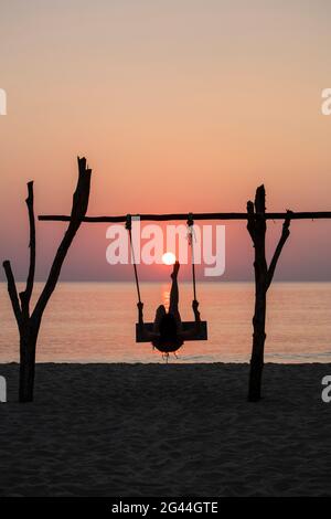 Silhouette einer jungen Frau, die bei Sonnenuntergang am Ong lang Beach auf Schaukel ist und vorgibt, die Sonne wie Fußball zu treten, Ong lang, Phu Quoc Island, Kien Giang, Stockfoto