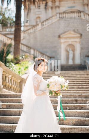 Schöne Braut in zartem Brautkleid mit Brautstrauß auf der Treppe der Geburtskirche der seligen Jungfrau Maria in P Stockfoto