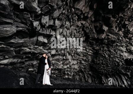 Hochzeit in Island. Hochzeitspaar unter einem Felsen aus Basaltsteinen. Braut und Bräutigam am schwarzen Strand von Vik. Stockfoto