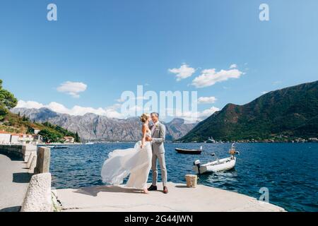 Braut und Bräutigam stehen umarmt am Pier in der Bucht von Kotor, Boote sind daneben Stockfoto