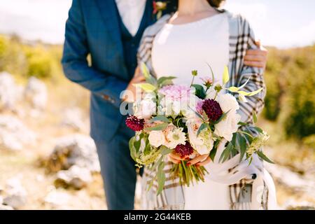 Der Bräutigam umarmt die Schultern der Braut in einem karierten Schal Mit einem schönen Blumenstrauß in ihren Händen Stockfoto