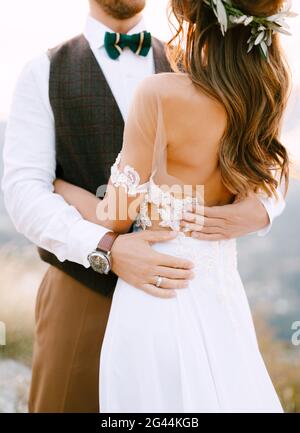 Halbportrait des Bräutigams, der die Braut umarmt und mit dem Rücken steht. Braut in einem schönen weißen Kleid und einem Kranz aus grünem Olivenzweig Stockfoto