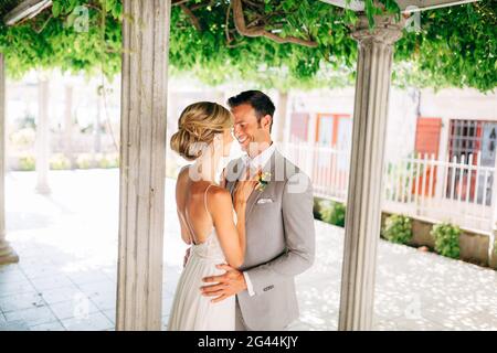 Braut und Bräutigam stehen zwischen den Säulen, umarmen und lächeln. Die Braut legte ihre Hände auf die Brust des Bräutigams. Stockfoto