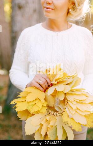 Frau in einem weißen Pullover hält einen gelben Kranz Der Herbst geht in ihren Händen Stockfoto