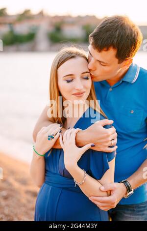Mann küsst lächelnde Schwangere sanft in einem blauen Kleid Sie umarmte ihre Schultern Stockfoto