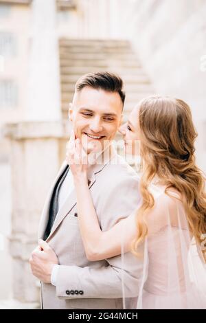 Die Braut hält ihre Handfläche auf dem Kinn des lächelnden Bräutigams Vor dem Hintergrund der Stufen eines alten Gebäudes In Bergamo Stockfoto