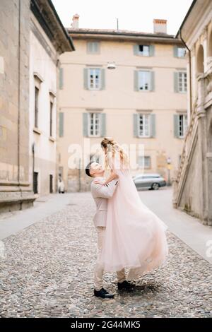 Bräutigam umkreist die Braut in seinen Armen und steht auf den Pflastersteinen in der Nähe des alten Gebäudes in Bergamo, Italien Stockfoto