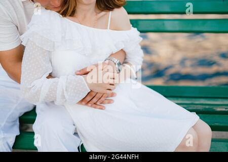 Ein Mann und eine Frau in weißen Kleidern sitzen auf einer grünen Bank, ein Mann umarmt sanft eine Frau von hinten, aus der Nähe Stockfoto