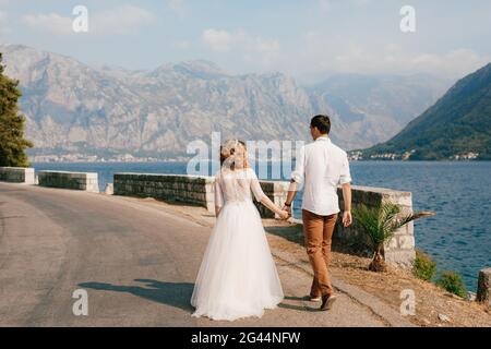 Braut und Bräutigam gehen Hand in Hand entlang der Straße entlang der Küste in der Bucht von Kotor bei Perast, Rückansicht Stockfoto