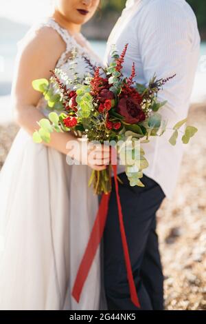 Braut und Bräutigam umarmen sich und halten einen Hochzeitsstrauß Am Kiesstrand am Meer Stockfoto