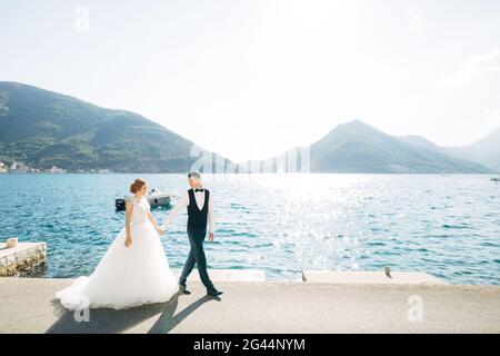 Die Braut und der Bräutigam gehen Hand in Hand auf der Straße am Meer, hinter ihnen liegen Berge und ein Boot Stockfoto
