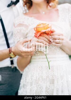 Der Bräutigam hält die Hand der Braut in einem weißen Spitzenkleid mit einer Rose in den Händen. Nahaufnahme Stockfoto