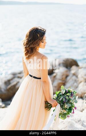 Schöne nachdenkliche Braut steht seitlich mit einem Bouquet von gedreht Blumen auf einem Felsen über dem Meer Stockfoto