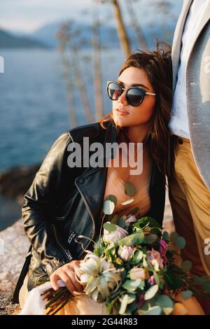 Braut in einer Lederjacke und Sonnenbrille mit einem Strauß Von Blumen sitzt auf einem Stein, der sich gegen den lehnt Bräutigam Stockfoto