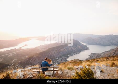 Die Braut und der Bräutigam sitzen und umarmen sich auf dem Holz Bank auf dem Gipfel des Mount Lovcen mit Blick auf die Bucht Von Kotor Stockfoto