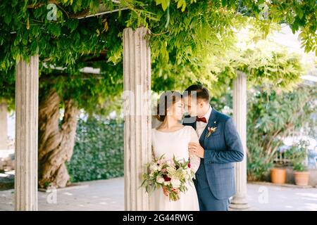 Lächelnder Bräutigam umarmt die Schultern der Braut vor dem Hintergrund von Säulen in einem grünen Garten Stockfoto