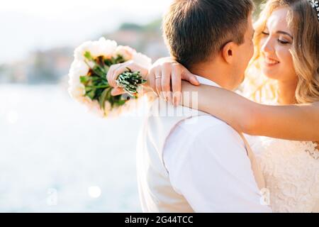 Die Braut umarmt den Bräutigam und hält einen Blumenstrauß in den Händen auf dem Pier in der Bucht von Kotor, Nahaufnahme Stockfoto