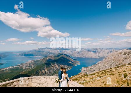 Bräutigam und Braut umarmen die Bucht von Kotor und blicken auf sie. Schöne Aussicht von den Bergen von Montenegro Stockfoto