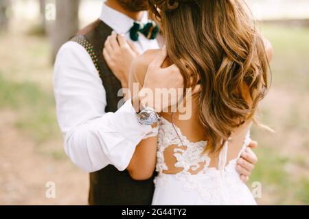 Der Bräutigam umarmt die Schultern der Braut in einem wunderschönen, bestickten weißen Kleid. Die Braut steht mit dem Rücken Stockfoto