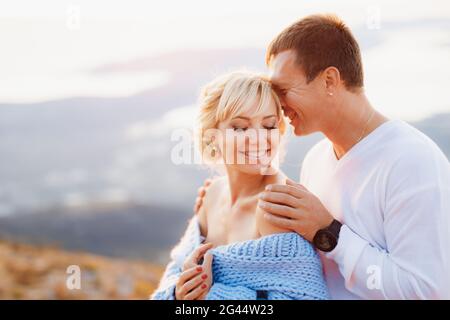 Lächelnder Mann umarmt lächelnde Frau in einem blauen Pullover an den Schultern auf dem Hintergrund der Berge. Nahaufnahme Stockfoto