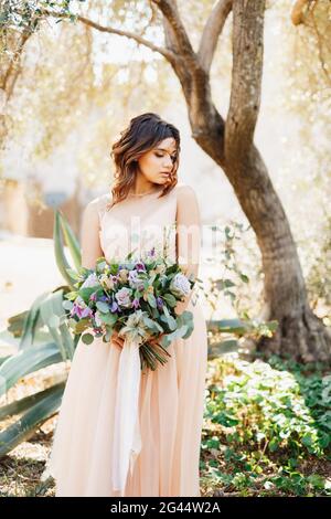 Niedliche Braut in einem schönen Kleid steht nachdenklich gegen ein Baumhintergrund mit einem wunderschönen Blumenstrauß in ihr Hände Stockfoto