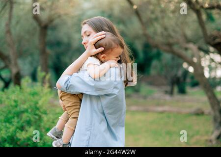 Mama hält ihren kleinen Sohn sanft in den Armen entgegen Der Hintergrund von Bäumen in einem Olivenhain Stockfoto