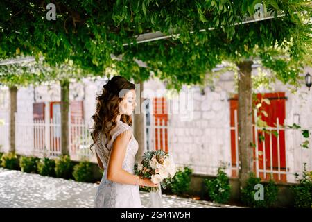 Schöne Braut in einem luxuriösen Kleid steht mit einem schönen Brautstrauß aus Rosen in einem grünen Garten an einem sonnigen Tag. Stockfoto