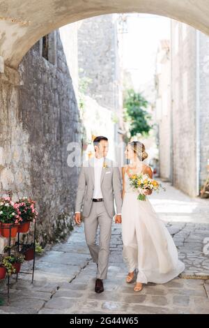 Braut und Bräutigam gehen mit einem Blumenstrauß Hand in Hand Hand entlang einer gemütlichen Straße der Altstadt unter Der Bogen und das Lächeln Stockfoto