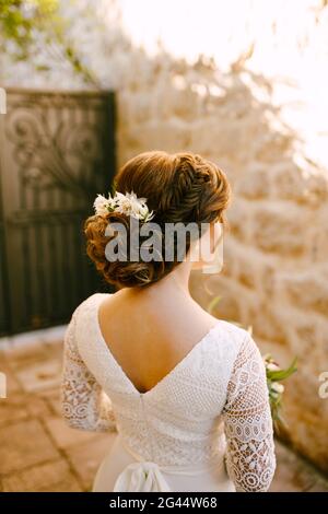 Stilvolle Braut steht zurück in einem weißen Spitzenkleid mit Eine luxuriöse Frisur und Blumen im Haar sieht an Die gegenüberliegende Wand