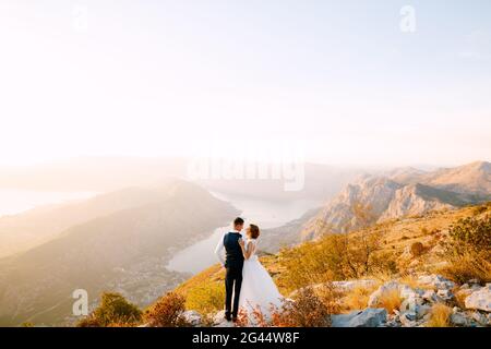 Die Braut und der Bräutigam stehen auf dem Gipfel des Mount Lovcen mit Blick auf die Bucht von Kotor, lächeln und umarmen sie zärtlich Stockfoto