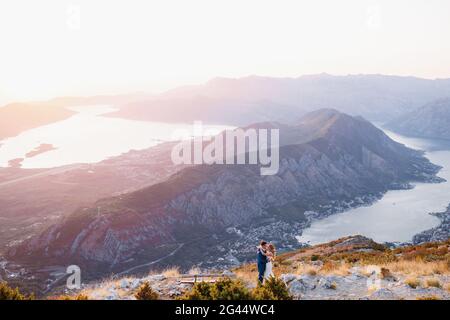 Die Braut und der Bräutigam stehen in der Nähe der Holzbank auf dem Gipfel des Mount Lovcen mit Blick auf die Bucht von Kotor, küssen und umarmen zehn Stockfoto