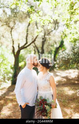 Die Braut und der Bräutigam stehen mit einem Strauß umarmt zwischen den Bäumen in einem Olivenhain, der Bräutigam küsst die Braut auf die Stirn Stockfoto