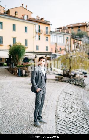 Der Bräutigam steht mit den Händen in den Taschen auf einer gepflasterten Straße vor dem Hintergrund uralter Villen. Comer See Stockfoto